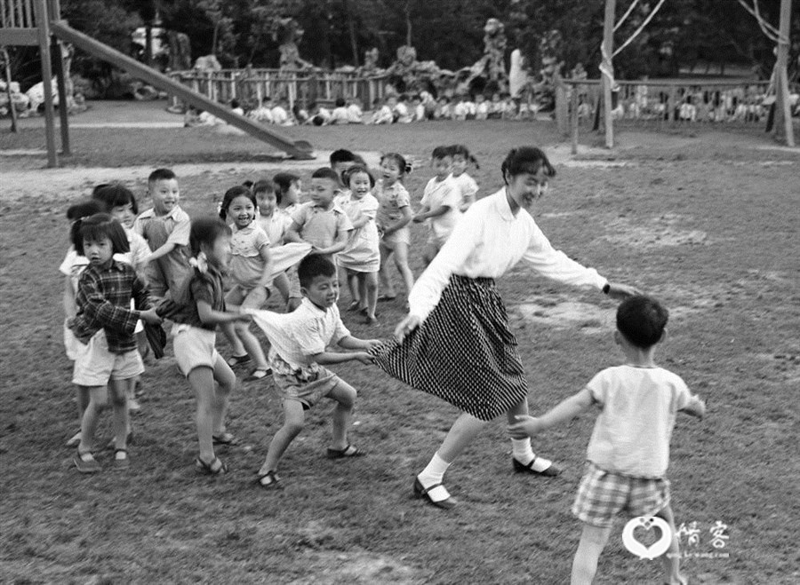 1959年，上海市一个幼儿园的草坪上，孩子们在阿姨的带领下作“老鹰捉小鸡”游戏。